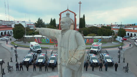 Aufschlussreiche-Drohnenaufnahme-Von-Sieben-Polizeiautos-Und-Zwei-Krankenwagen-Mit-Bewaffneten-Beamten-Der-Polizei,-Die-Vor-Ihren-Autos-Und-Hinter-Einer-Statue-Von-Präsident-Benito-Juárez-Posieren