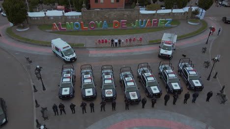 Toma-De-Seguimiento-A-Gran-Altura-De-7-Camionetas-Policiales-Y-2-Ambulancias-Con-Oficiales-Armados-Del-Departamento-De-Policía-Alineados-Y-Posando-Frente-A-Sus-Autos-Y-Detrás-De-Una-Estatua-Del-Presidente-Benito-Juárez.