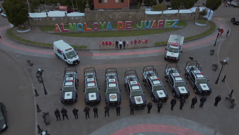 High-altitude-tracking-shot-of-7-police-pick-ups-and-2-ambulances-with-armed-officers-of-the-Police-Department-lined-up-and-posing-in-front-of-their-cars-and-behind-a-statue-of-President-Benito-Juarez