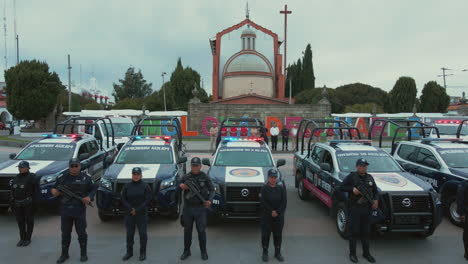 Vertikaler-Schwenk-Von-7-Polizei-Pickups-Und-2-Krankenwagen-Mit-Bewaffneten-Beamten-Der-Polizei,-Die-Vor-Ihren-Autos-Aufgereiht-Und-Posieren,-Mit-Einer-Kirche-Im-Hintergrund