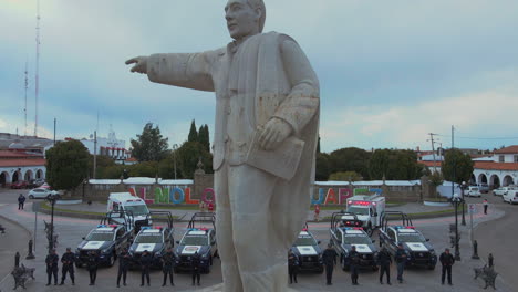 Aufschlussreiche-Drohnenaufnahme-Von-Sieben-Polizeiautos-Und-Zwei-Krankenwagen-Mit-Bewaffneten-Beamten-Der-Polizei,-Die-Vor-Ihren-Autos-Und-Hinter-Einer-Statue-Von-Präsident-Benito-Juárez-Posieren