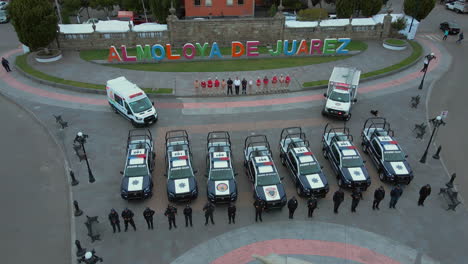 Tracking-shot-of-7-police-pick-ups-and-2-ambulances-with-armed-officers-of-the-Police-Department-lined-up-and-posing-in-front-of-their-cars-and-behind-a-statue-of-President-Benito-Juarez
