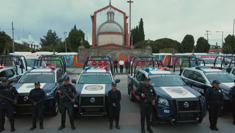 Vertical-pan-of-7-police-pick-ups-and-2-ambulances-with-armed-officers-of-the-Police-Department-lined-up-and-posing-in-front-of-their-cars,-with-a-church-in-the-background