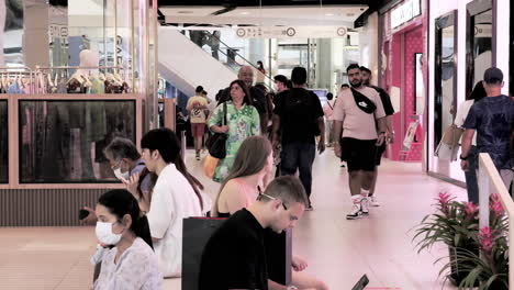 Touristen-Und-Käufer-Sind-Mit-Einem-Schaufensterbummel-Nach-Designer-Modemarken-Beschäftigt,-Während-Einige-Einfach-Nur-Sitzen-Und-Ihre-Mobiltelefone-In-Einem-Einkaufszentrum-In-Bangkok,-Thailand,-Checken