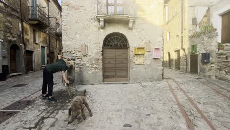 Man-and-dog-drinking-at-street-fountain-during-grape-harvest-festival-of-medieval-Penna-in-Teverina-town-with-decorated-houses-and-streets-in-Italy