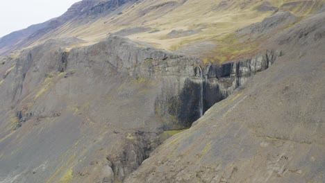 Aerial-approaching-shot-of-waterfall-in-rocky-and-volcanic-mountain-landscape-Iceland