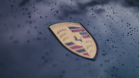 Primer-Plano-Del-Emblema-De-Porsche-En-El-Capó-Negro-Del-Coche-Mojado