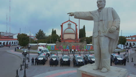 Vertikaler-Schwenk-Von-7-Polizei-Pick-ups-Und-2-Krankenwagen-Mit-Bewaffneten-Beamten-Der-Polizei,-Die-Vor-Ihren-Autos-Und-Hinter-Einer-Statue-Von-Präsident-Benito-Juárez-Aufgereiht-Sind-Und-Posieren