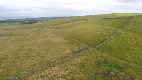 Slow-moving-reversing-drone-footage-over-the-hills-of-West-Yorkshire-countryside,-farmland-and-moorland-flying-parallel-to-a-farmer's-track-and-over-a-dry-stone-wall-with-towns-in-the-far-distance