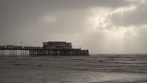 Sunbeams-burst-through-fast-moving-storm-clouds-as-they-pass-over-dark-and-threatening-choppy-waves-rolling-in-around-a-pleasure-pier-before-storm-Ciarán-makes-landfall