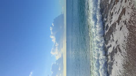 Vertikales-Bild-Eines-Ruhigen-Strandes-Ohne-Menschen,-Weißer-Schaum,-Kleine-Steine,-Kopienraum-Am-Blauen-Himmel