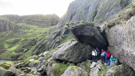 Hillwalkers-sheltering-from-winter-rain-under-large-rocks-and-high-cliffs-at-Coumshingaun-Lake-Comeragh-Mountains-on-a-cols-winter-day