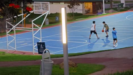 Un-Adolescente-Lo-Intenta,-Pero-Falla-Y-Se-Cae-Mientras-Juega-Un-Partido-De-Baloncesto-Al-Atardecer-En-Una-Cancha-Pública-Con-Dos-Jugadores,-Un-Contenedor-De-Basura-Y-Un-Poste-De-Luz-En-Un-Parque-En-Buen-Estado.