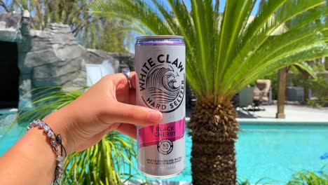 Holding-a-White-Claw-hard-seltzer-black-cherry-can-at-a-luxury-Las-Vegas-villa-mansion,-fun-summer-party-vibe-with-swimming-pool-view-and-palm-trees,-4K-shot