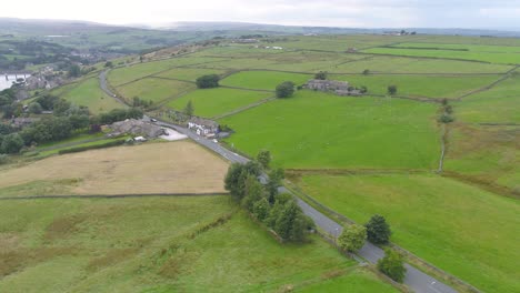 Strafing-drone-footage-descending-down-a-valley-in-Yorkshire-countryside-including-village-houses,-farms,-stone-walls,-country-roads-and-moving-cars,-farmer's-fields-and-moorland-hills-in-the-distance