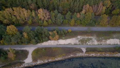 Aerial-shot-of-a-quiet-road-between-a-forest-with-autumn-foliage-and-a-rocky-shoreline