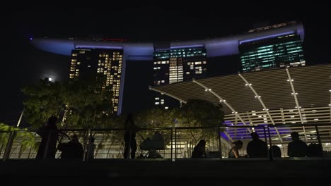 Looking-Up-At-Illuminated-Marina-Bay-Sands-Hotel-In-Singapore-At-Night-From-The-Boardwalk-With-Silhouette-Of-People-Beside-Railings