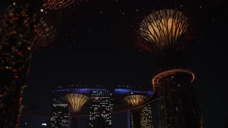 Mirando-Hacia-Los-Hermosos-Superárboles-Iluminados-En-Los-Jardines-Junto-A-La-Bahía-Por-La-Noche-En-Singapur-Con-El-Hotel-Marina-Bay-Sands-Al-Fondo