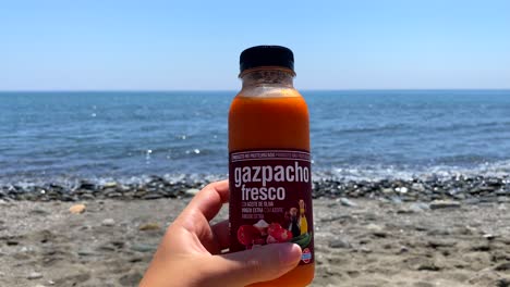 Halten-Sie-Eine-Frische-Traditionelle-Gazpacho-Miniflasche-Aus-Dem-Spanischen-Mercadona-Supermarkt-Am-Sonnigen-Strand-Von-Estepona,-Spanien,-Mit-Meerblick,-Kalte-Suppe-Für-Unterwegs,-Statische-4K-Aufnahme