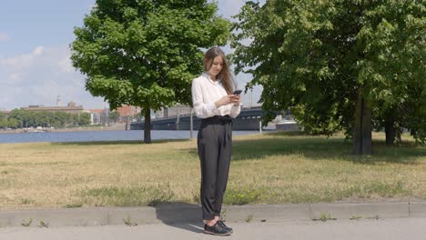 Woman-standing-still-on-the-sidewalk-using-her-smartphone-under-the-sun-and-looks-at-the-camera,-static-wide