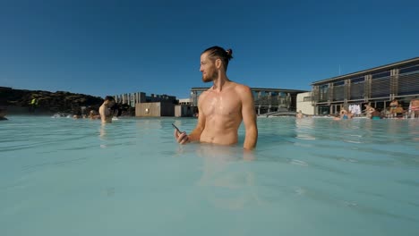 Hombre-Caucásico-Disfrutando-De-Un-Lago-Termal-Mientras-Sostiene-Su-Teléfono-En-Islandia,-Atracción-Turística-Laguna-Azul