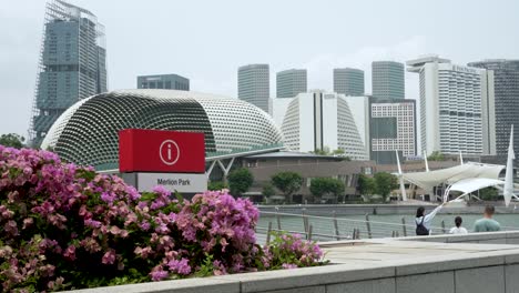 Merlion-Park-Schild-Mit-Esplanade---Theater-An-Der-Bucht-Im-Hintergrund-In-Singapur-Und-Touristen,-Die-Auf-Der-Jubiläumsbrücke-Stehen
