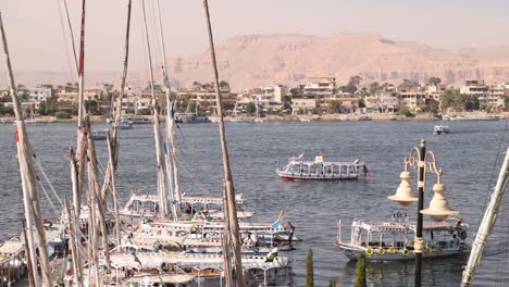 Schwenkaufnahme-Von-Booten,-Die-Entlang-Des-Nils-Treiben,-Mit-Bergen-Im-Hintergrund-Und-Segelbooten-Im-Vordergrund-In-Luxor,-Ägypten