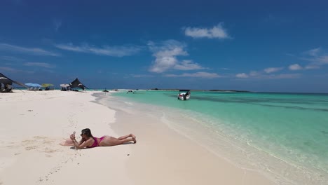Woman-lying-face-down-on-white-sand-beach-taking-photos-with-phone,-azure-caribbean-sea-background