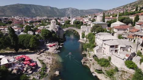 Aerial-view-circling-the-iconic-Stari-Most-with-motor-boat-travelling-towards-the-old-arched-bridge-in-Mostar,-Bosnia