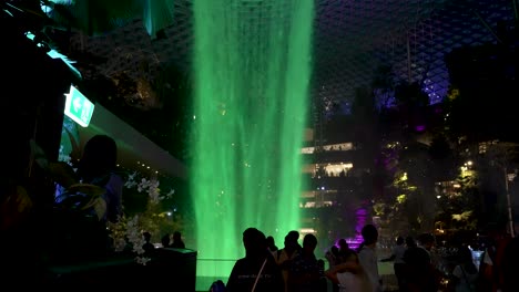 Cascada-Interior-Iluminada-De-Color-Verde-Brillante-En-El-Aeropuerto-Jewel-Changi-Con-Visitantes-Y-Gente-Caminando-Tomando-Fotos-Al-Lado