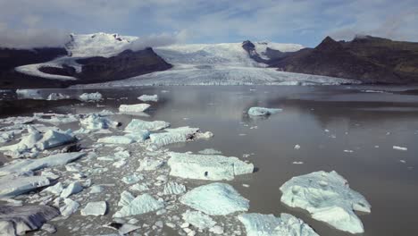 Vuelo-Aéreo-Sobre-Dramáticos-Icebergs-Flotando-En-El-Agua,-Lago-Jokulsarlon,-Paisaje-Nevado-De-Invierno,-Glaciar-A-Distancia,-Islandia