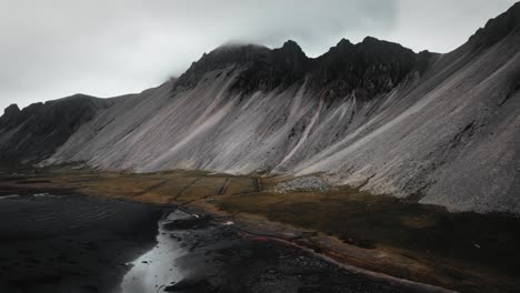 Playa-Aérea-De-Arena-Negra-Stokksnes,-Montañas-Volcánicas-Oscuras,-Con-Hierba-Verde-Naranja,-Paisaje-Oscuro-Y-Nublado,-Islandia