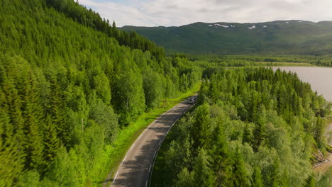 Drone-follows-campervan-driving-along-winding-road-in-mountains-past-lake,-Norway