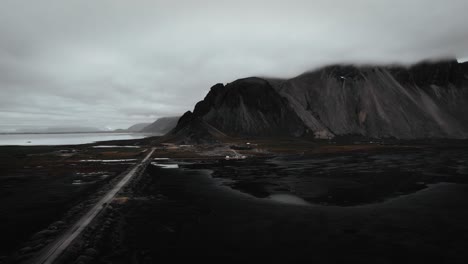 Aerial-black-sand-beach-stokksnes,-volcanic-dark-mountains-in-distance,-dark-moody-cloudy-seagulls-flying,-Iceland