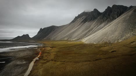 Aerial-black-sand-beach-stokksnes,-volcanic-dark-mountains,-with-green-orange-grass,-dark-moody-cloudy-scenery-Iceland