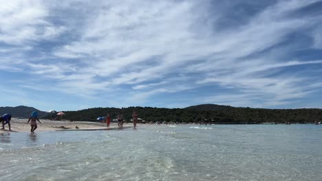 Low-angle-water-surface-view-of-people-bathing-and-sunbathing-on-white-sand-of-Lotu-beach-in-summer-season,-Corsica-island-in-France