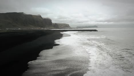 Cinematic-aerial-flying-over-black-sand-beach-ocean-scenery,-moody-dark-scenery,-town-Vik,-Iceland