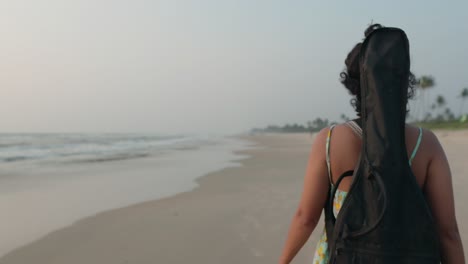 A-young-Indian-musician,-with-her-guitar-case-on-her-back,-immerses-herself-in-the-serene-beauty-of-the-beach,-creating-a-harmonious-fusion-of-music-and-nature