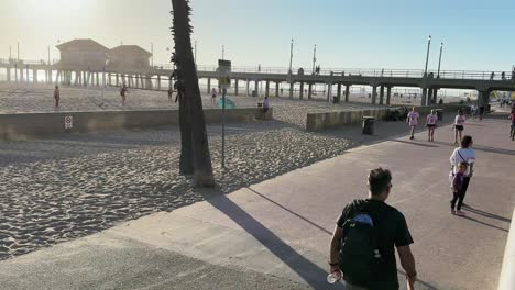 People-cycling,-playing,-walking-on-promenade-near-sandy-beach-and-pier-in-Huntington-Beach,-California