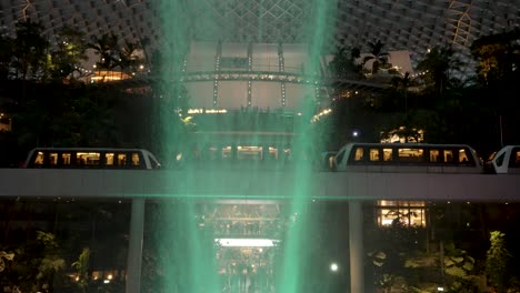 Skytrain's-Passing-Each-Other-Beside-Indoor-Waterfall-Illuminated-By-Green-Light-At-Jewel-Changi-Airport