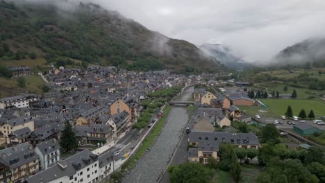 A-breathtaking-drone-shot-captures-the-view-of-a-charming-Pyrenees-mountain-town-on-the-Spanish-side,-perched-above-a-tranquil-riverbed,-enveloped-in-cozy-charm-on-a-cold,-cloudy-day
