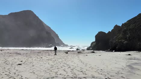 Eine-Person,-Die-An-Einem-Sandstrand-Mit-Felsen-Und-Wellen-Und-Vielen-Schritten-Im-Sand-Spaziert