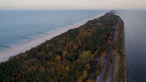 Aerial-view-of-a-coastal-road-with-adjacent-beach-and-sea-on-one-side-and-autumnal-forest-on-the-other-during-dusk---Kuznica-Poland
