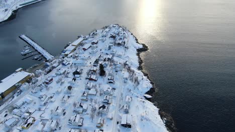 Drone-view-on-the-Tromso-mountains-in-winter-full-of-snow-circling-around-Husoy,-a-small-town-on-an-island-surrounded-by-the-sea