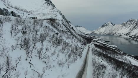 Drone-view-in-Tromso-area-in-winter-lifting-from-the-ground-showing-a-fjord-surrounded-by-white-mountains-in-Norway