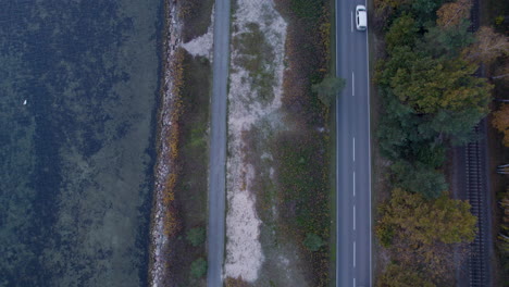 Overhead-shot-of-a-two-lane-road-with-vehicles-between-a-sandy-area-and-a-forest-with-autumn-foliage