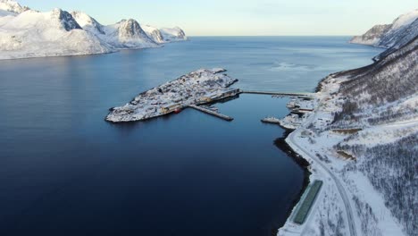 Drone-view-on-the-Tromso-mountains-in-winter-full-of-snow-showing-Husoy-a-small-town-on-an-island-surrounded-by-the-sea-and-its-small-port-with-flying-seagulls