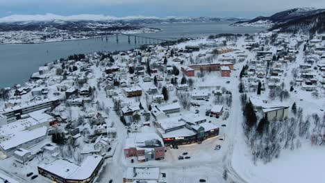 Drone-view-in-Tromso-of-Finnsnes,-a-small-town-full-of-snow-and-mountains-in-the-horizon-in-Norway-and-a-bridge