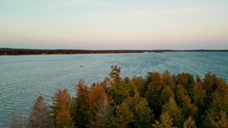 Aerial-view-over-changing-leaves-lakeside-with-canoe-crossing-lake,-Les-Cheneaux-Islands,-Michigan