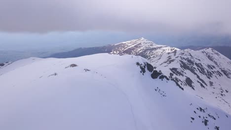 Ascending-aerial-view-of-climber-on-summit-of-snow-covered-mountain-peak-and-footprints-in-snow-landscape-along-ridge